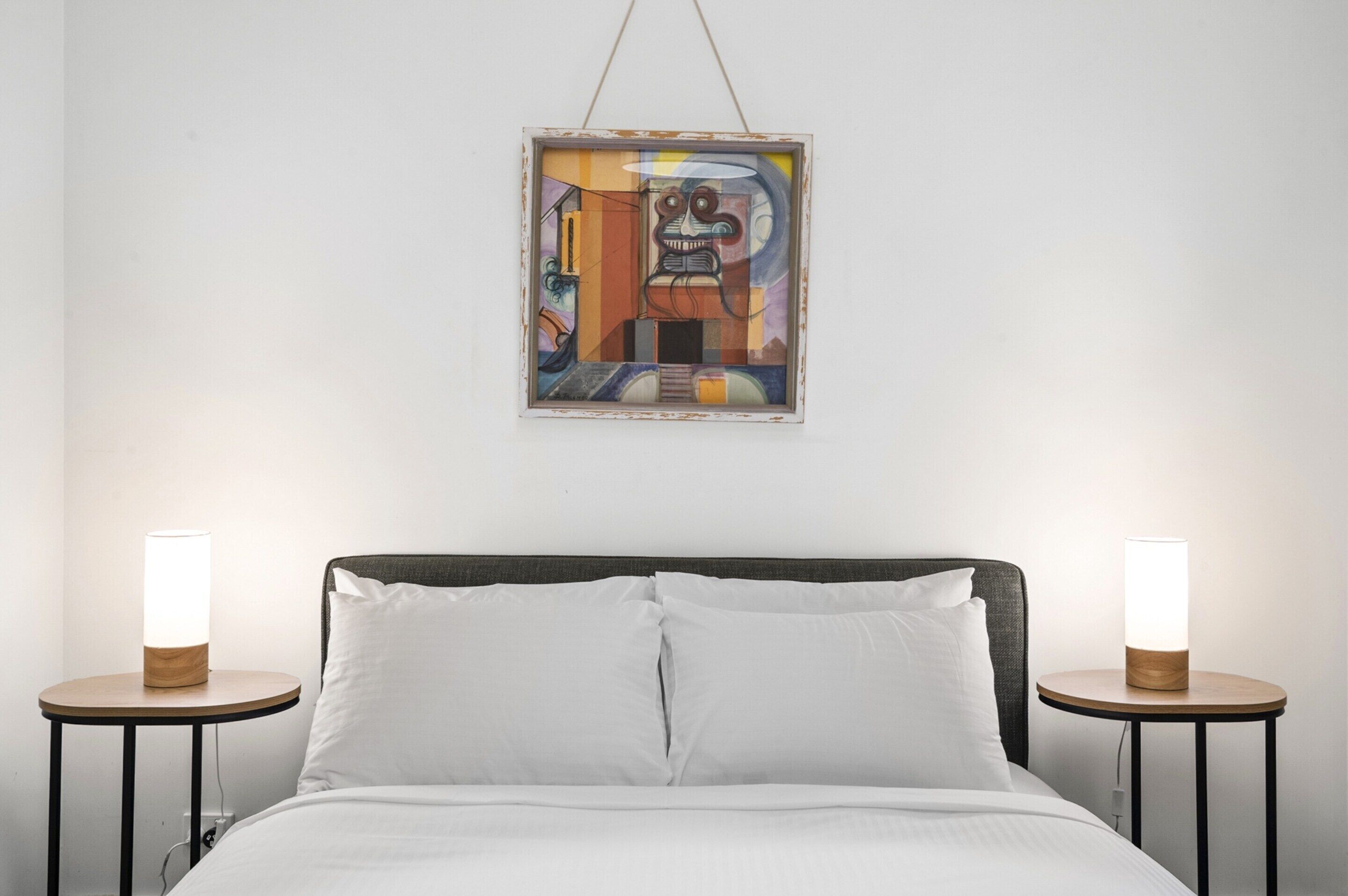 Una cama con ropa blanca, dos mesitas de noche con lámparas y un cuadro abstracto enmarcado en la pared.