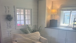 Ett vardagsrum med en beige soffa, ett litet vitt bord, ett fönster med utsikt och ett skåp med en lampa.
