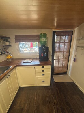 Una cocina con piso de madera, dispensador de agua, ventana con persianas y puerta con felpudo.
