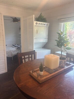 Ein Zimmer mit Esstisch aus Holz, einer integrierten Waschmaschine und einem Trockner, einer kleinen Pflanze und einem Fenster mit Blick auf Grün.