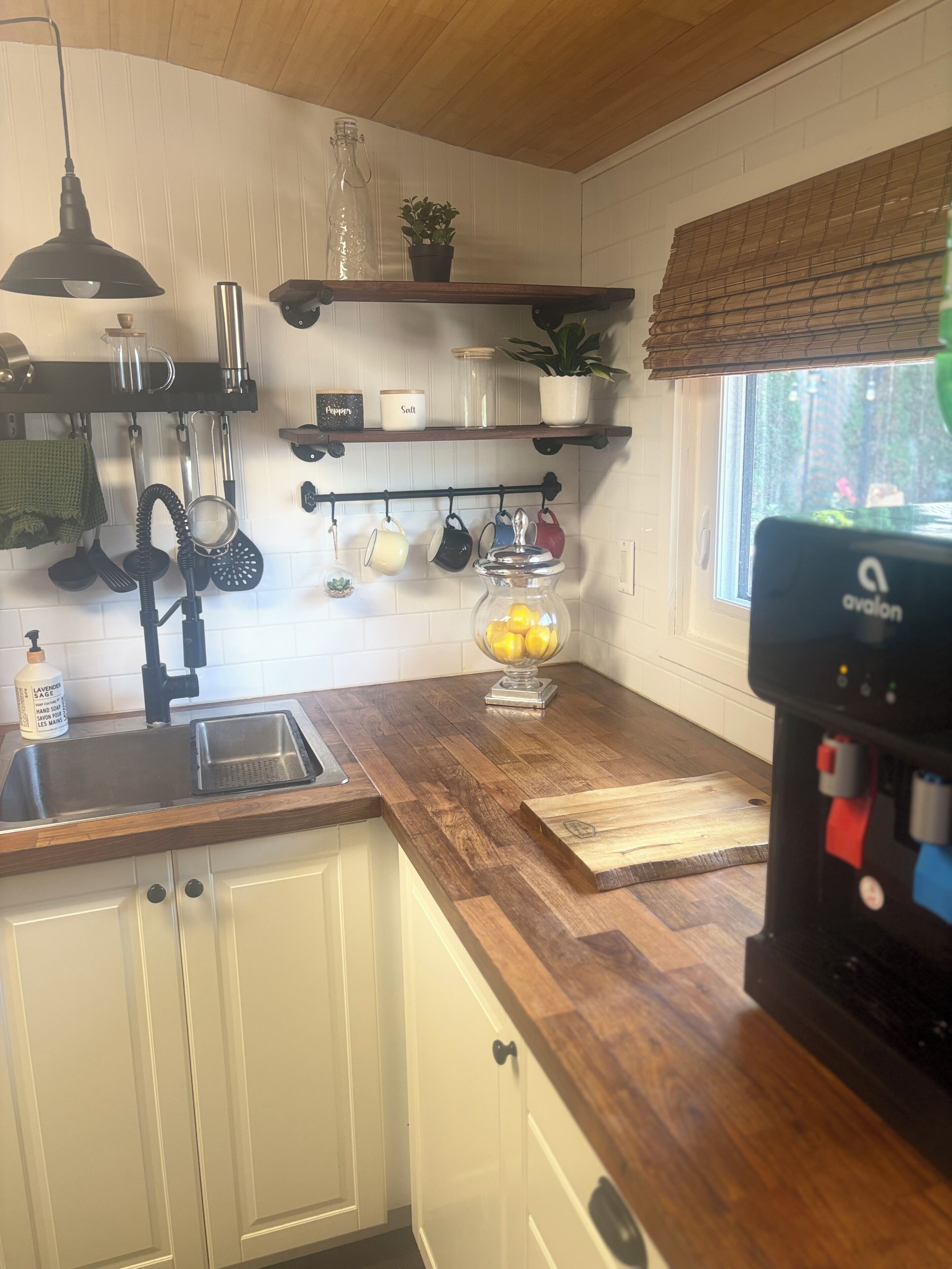 Une cuisine dotée d’un plan de travail en bois, d’un évier, d’une étagère à ustensiles suspendue et d’une fenêtre donnant sur un paysage verdoyant.