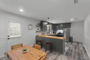 A compact kitchen with a central island, a dining area with a wooden table, and a small bar stool. - The Paddle House 3/2 Walking Distance To New Pickleball Courts (Dothan)