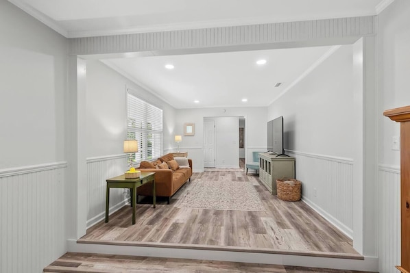 A hallway with a wooden floor, a brown sofa, a small table with a lamp, and a television on a cabinet. - The Paddle House 3/2 Walking Distance To New Pickleball Courts (Dothan)