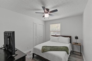 Un dormitorio con cama, ventilador de techo, televisor, mesita de noche y ventana con persianas.