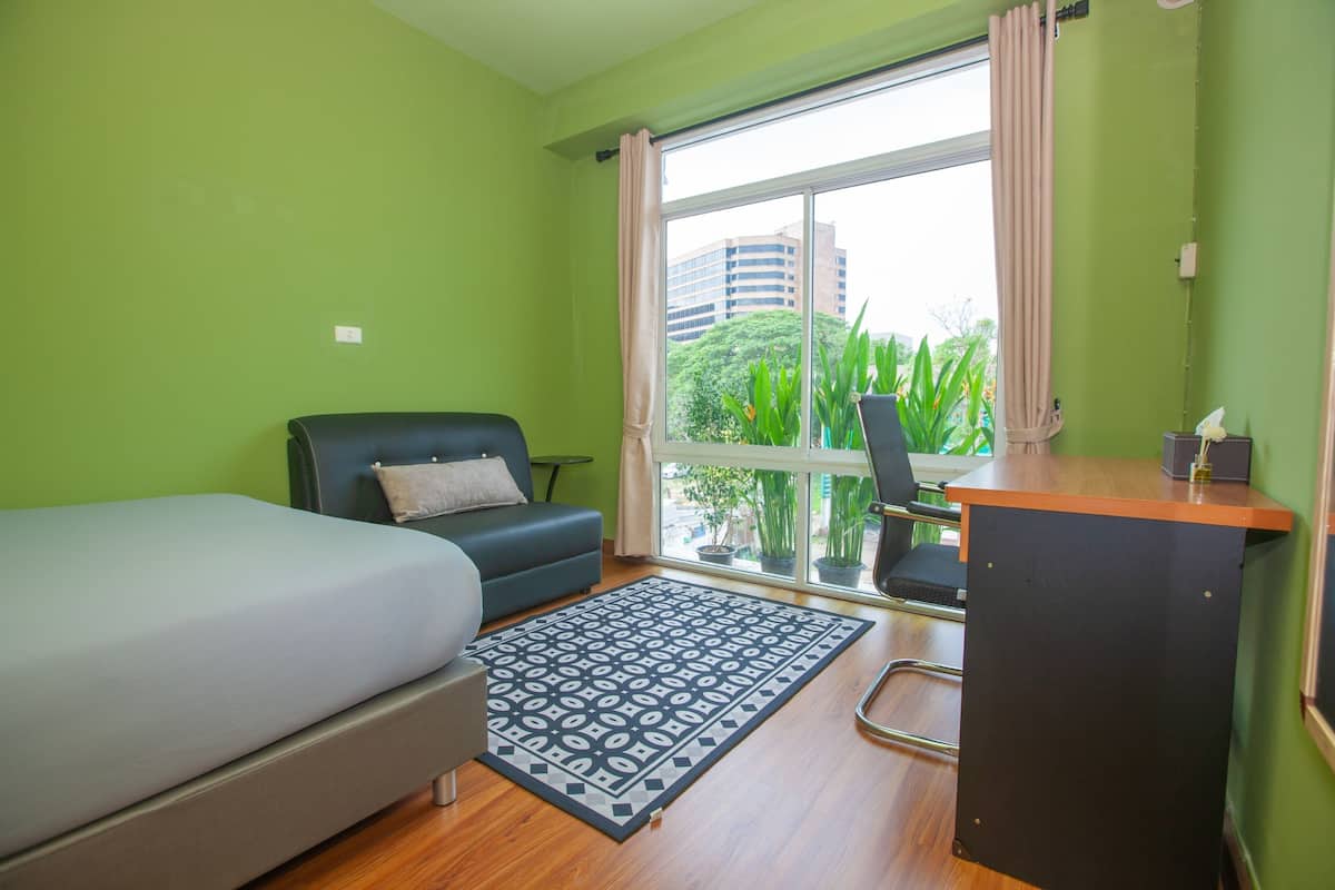 A room with a bed, a desk, a chair, a sofa, and a window with a view of buildings and greenery.