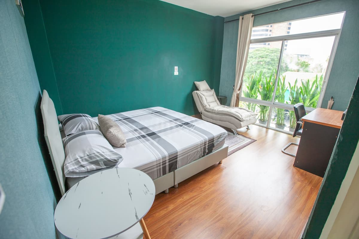 A bedroom with a bed, a chair, a small table, and a view of greenery outside.