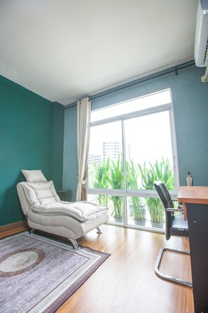 A room with a massage chair, a desk, a large window with a view of greenery, and a patterned rug.