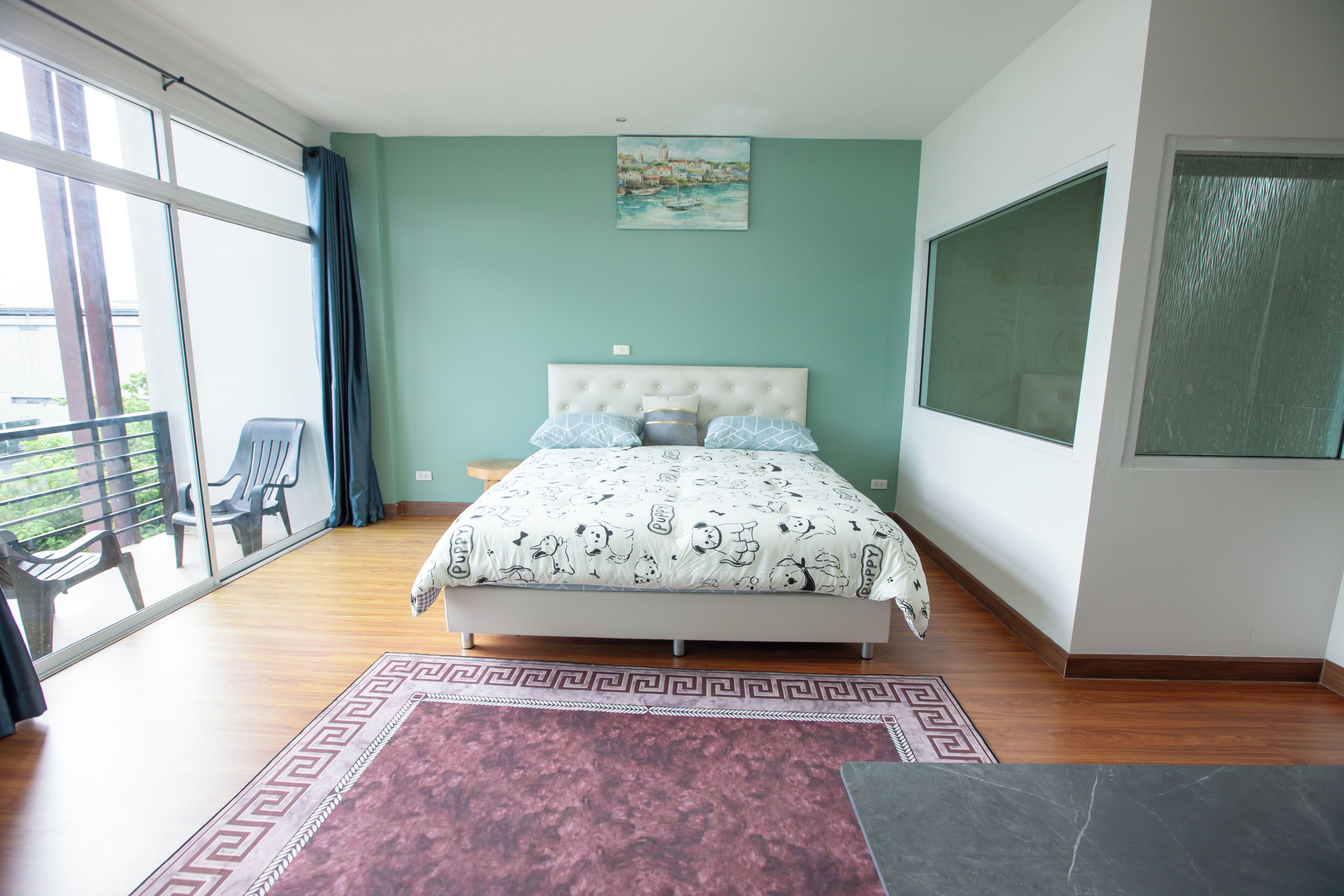 A bedroom with a bed, a large window, a balcony with chairs, and a painting on the wall.