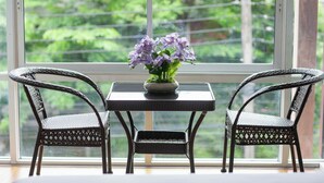 Una habitación con ventana, dos sillas de mimbre, una mesita y un florero con flores.