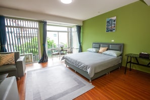A bedroom with a bed, a sofa, a small table, a chair, and a view through a large window.