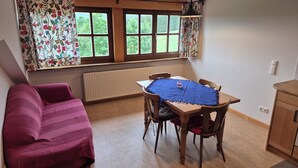 Ein Zimmer mit Bett, Esstisch mit Stühlen, einem Fenster mit Vorhängen, einem Heizkörper und einem Schrank.
