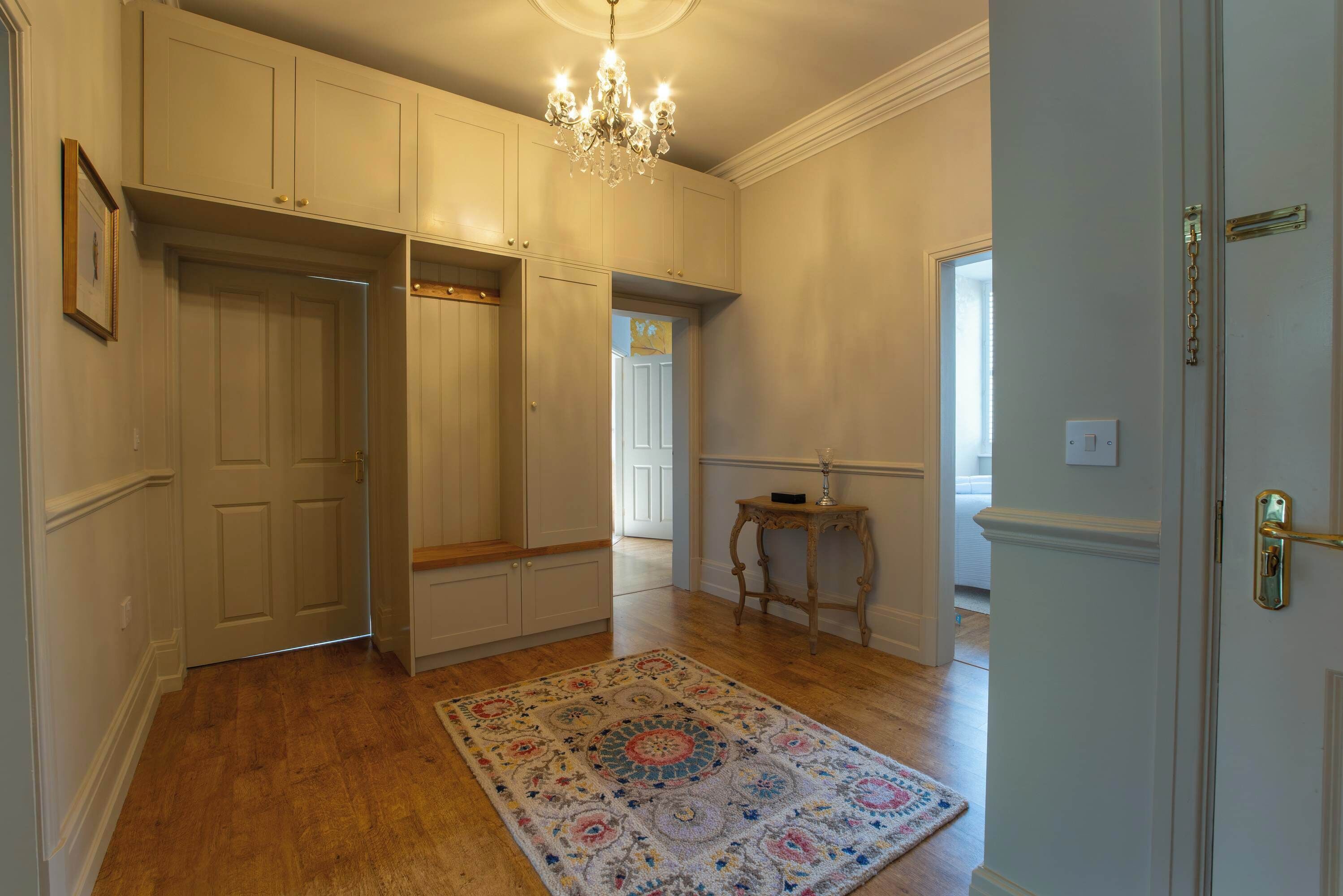 Un couloir doté d’un plancher en bois, d’un tapis à motifs, d’un lustre et d’une porte avec une poignée dorée.