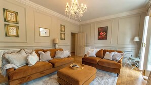 Ein Wohnzimmer mit zwei braunen Sofas, einem passenden Hocker, einer Kandelaber und gerahmten Kunstwerken an den Wänden. Eine Tür führt in einen anderen Raum.
