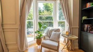 Ein Zimmer mit einem großen Erkerfenster, einem Sessel, einem kleinen Tisch und einem Bücherregal.