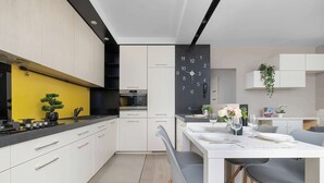 Eine moderne Küche mit weißen Schränken, einer gelben Akzentwand und einem Essbereich mit weißem Tisch und grauen Stühlen.