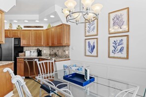 A modern kitchen with wooden cabinets, stainless steel appliances, a glass dining table, and framed botanical artwork on the wall. - Summer Breeze, Beach, Beach Bar, Sky Bridge, Pool (Miramar Beach)