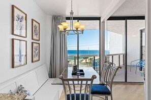 A balcony with a view of the beach, a dining area with a table and chairs, and framed artwork on the wall. - "Summer Breeze" Beach Access, Beach Bar, Sky Bridge, Pool, Hot Tubs, Bonfires (Miramar Beach)