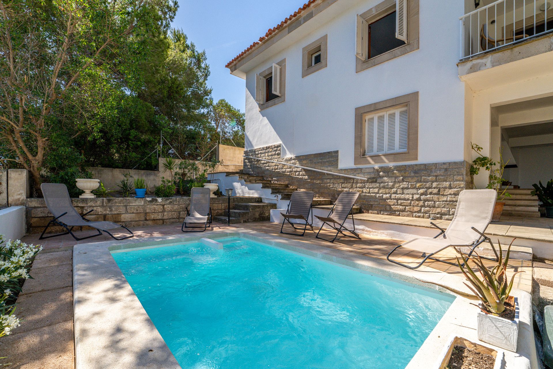 A pool area with lounge chairs, a stone wall, and a building with windows and shutters.