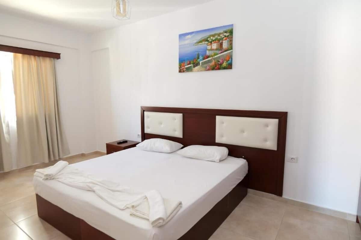 Una habitació d'hotel amb un llit gran, una taula de nit, un quadre a la paret i una finestra amb cortines.