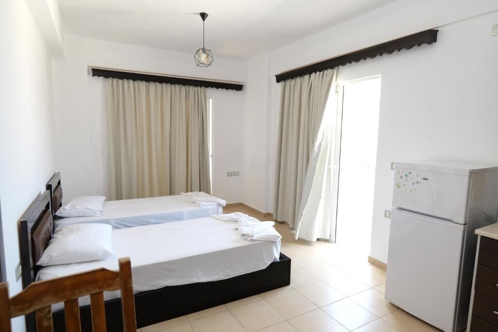 Habitación de hotel con dos camas, una silla de madera, un refrigerador y cortinas en las ventanas.