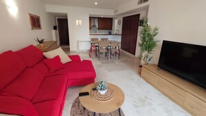 A living room with a red sofa, a coffee table with a plant, a TV, and a kitchen in the background. - Apartment in La Recoleta Punta Prima, Communal pools and steps away from the sea (Punta Prima)