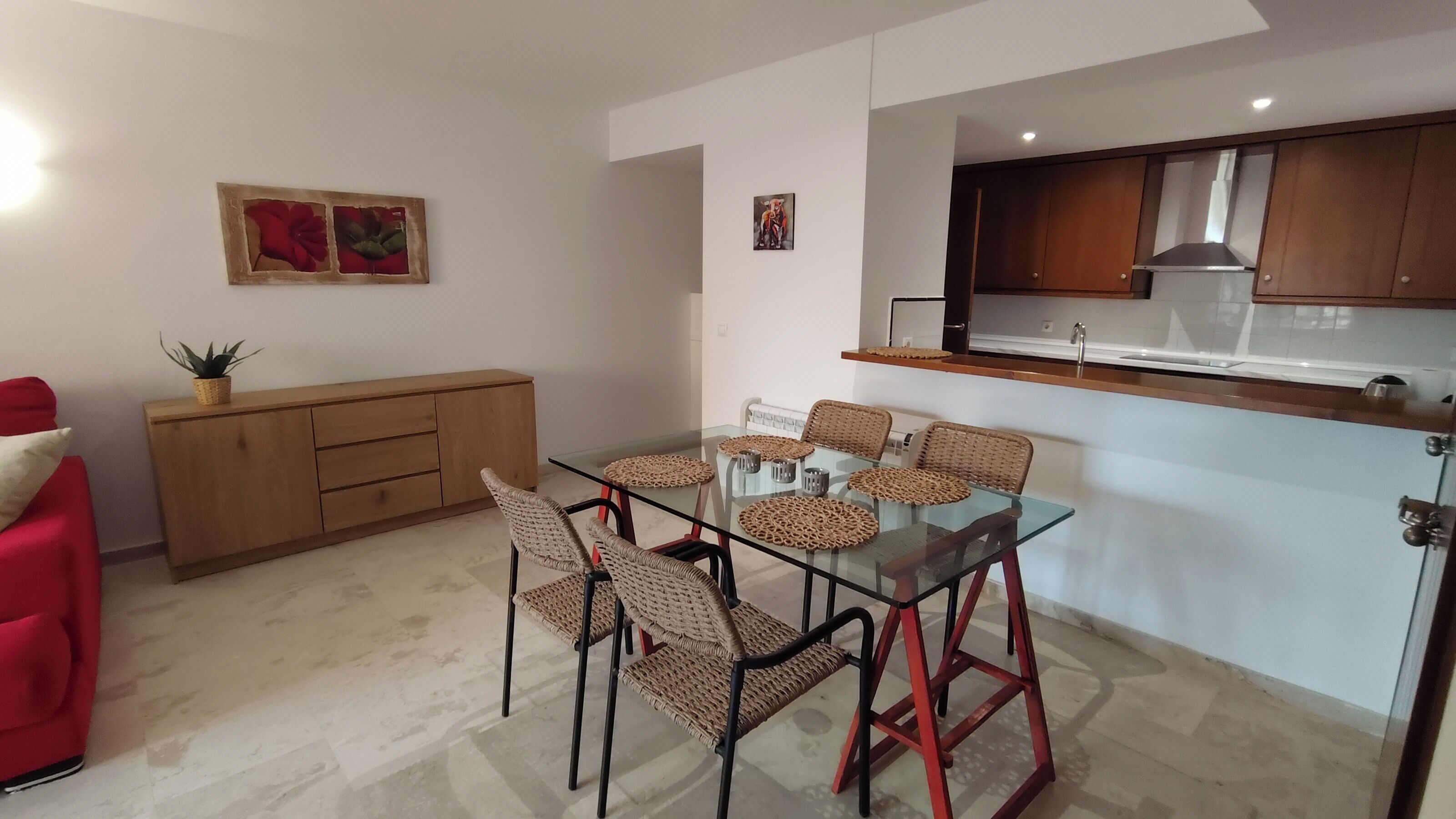 Een moderne keuken met houten kastjes, een glazen eettafel met geweven stoelen en een rode bank.