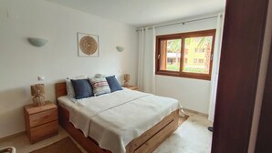 A bedroom with a wooden bed, a bedside table, a lamp, and a window with a view of buildings. - Apartment in La Recoleta Punta Prima, Communal pools and steps away from the sea (Punta Prima)