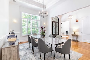 Uma sala de jantar com uma mesa de mármore, seis cadeiras, um vaso com flores, um candeeiro e uma janela grande com persianas.