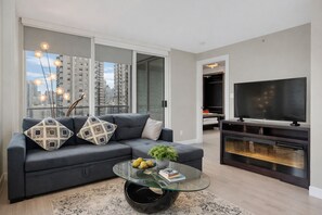 A modern living room with a grey sofa, patterned cushions, a glass coffee table with books and fruit, a flat-screen TV, and a view of high-rise buildings.