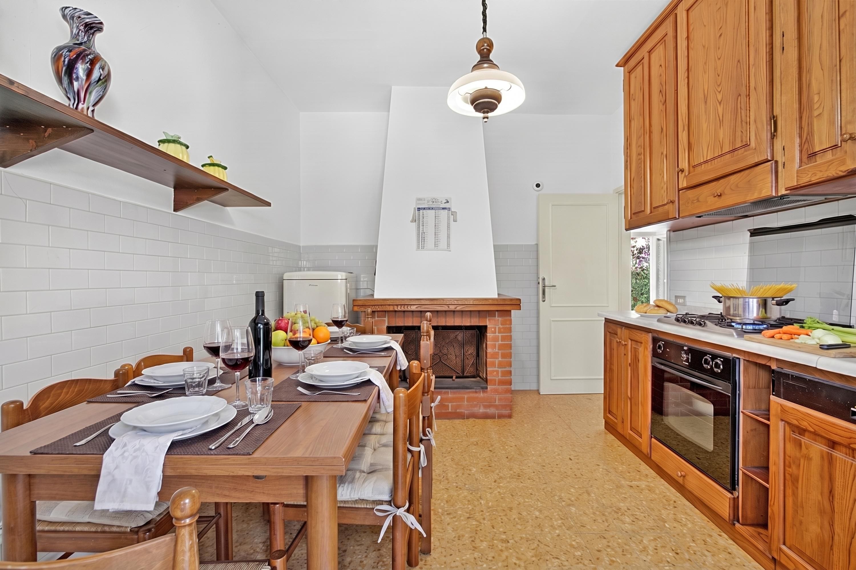 Een keuken met een eettafel gedekt voor vier personen, houten keukenkastjes, een haard van baksteen en een hanglamp.