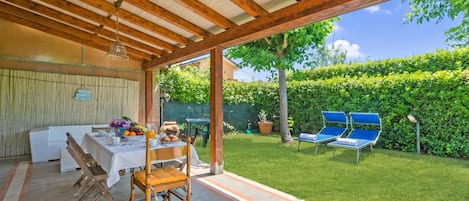 Une terrasse extérieure avec une table dressée pour les repas, un espace salon abrité et une vue sur un jardin bien entretenu.