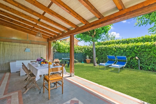 An outdoor patio with a table set for dining, a covered seating area, and a view of a well-maintained garden. - Casa Colombo With Garden In Forte Dei Marmi (Forte dei Marmi)