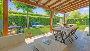 Une terrasse couverte avec une table dressée pour les repas, des sièges d’extérieur et une vue sur un jardin bien entretenu.