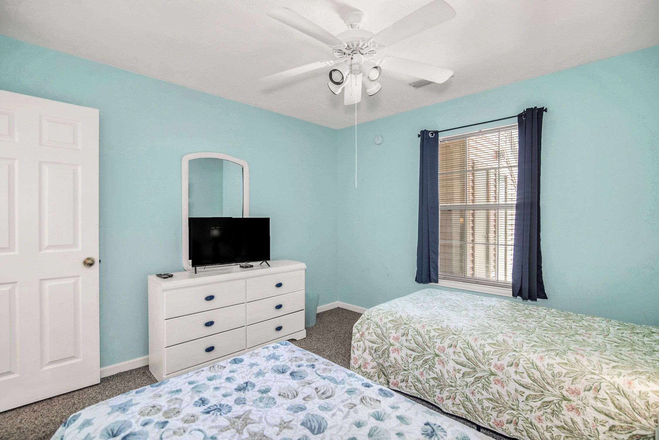 Ein Schlafzimmer mit weißem Schminktisch, einem Bett mit Blumenmuster auf der Bettwäsche, einem Deckenventilator und einem Fenster mit Vorhängen.