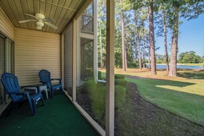 A covered porch with a ceiling fan, two Adirondack chairs, a small table, and a view of a lake surrounded by trees. - TB 24 three bedroom True Blue Condo (Pawleys Island)