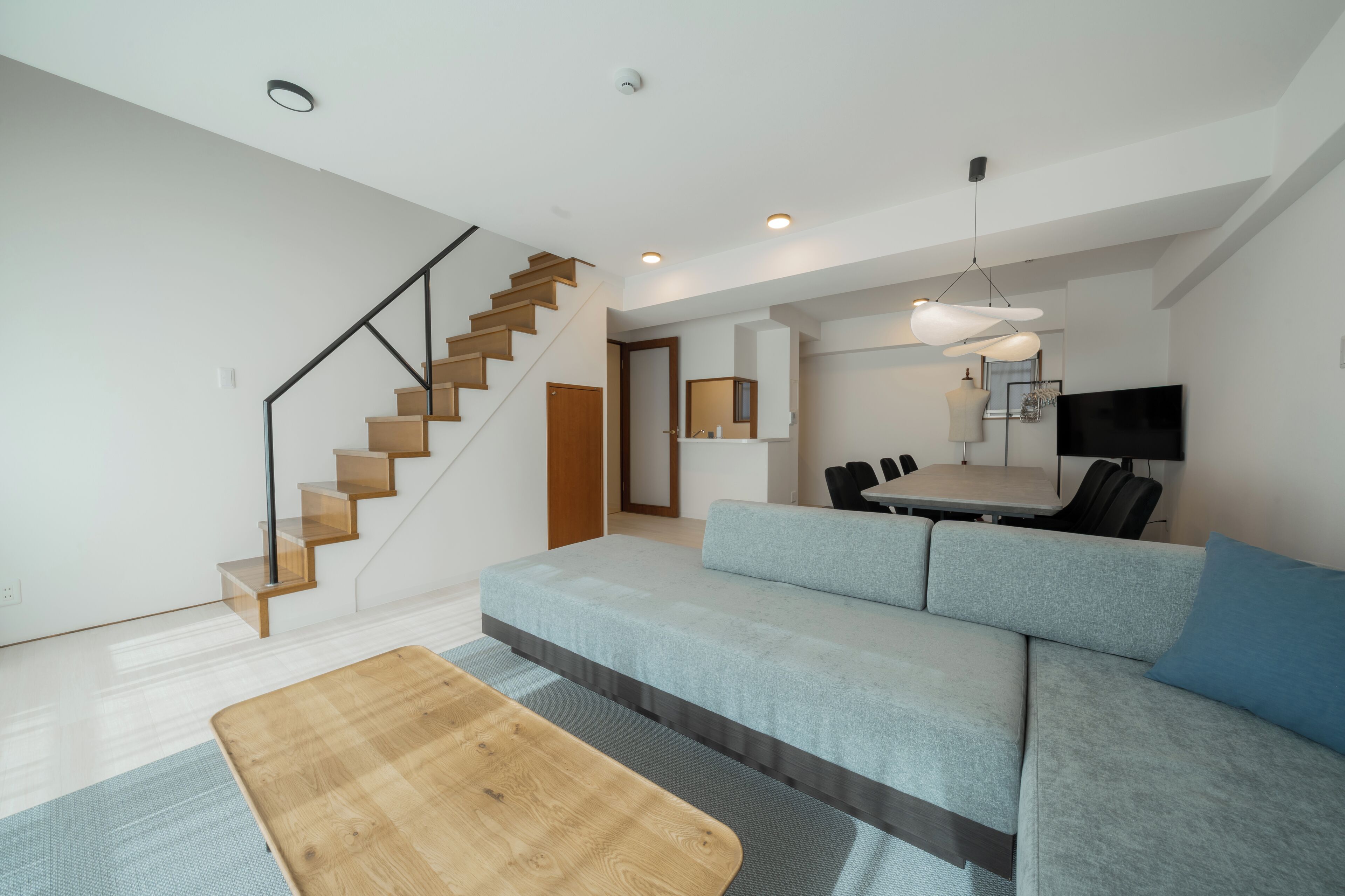 A modern living room with a staircase, a sofa, a dining table with chairs, and a flat-screen TV.
