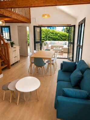 A modern interior with a wooden staircase, a dining area with a table and chairs, a blue sofa, and a view of greenery outside.