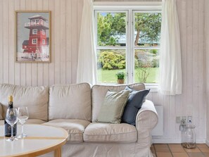 Ein beiges Sofa mit Kissen, ein Tisch mit Flasche und Gläsern, ein Fenster mit Vorhängen und ein gerahmtes Bild an der Wand.