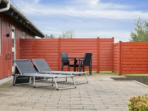 Eine Terrasse mit zwei Liegestühlen und einem Tisch, ein roter Zaun und ein Gebäude mit einem Metalldach.