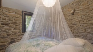 A room with a stone wall, a bed with a floral bedspread, and a canopy bed with a white net. - Charming Stone Cottage for Rent in a Picturesque Pyrenean Village (Glorianes)