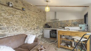 A stone-walled room with a sofa, a wooden table, a kitchen area with a stove, and a hanging light fixture. - Charming Stone Cottage for Rent in a Picturesque Pyrenean Village (Glorianes)