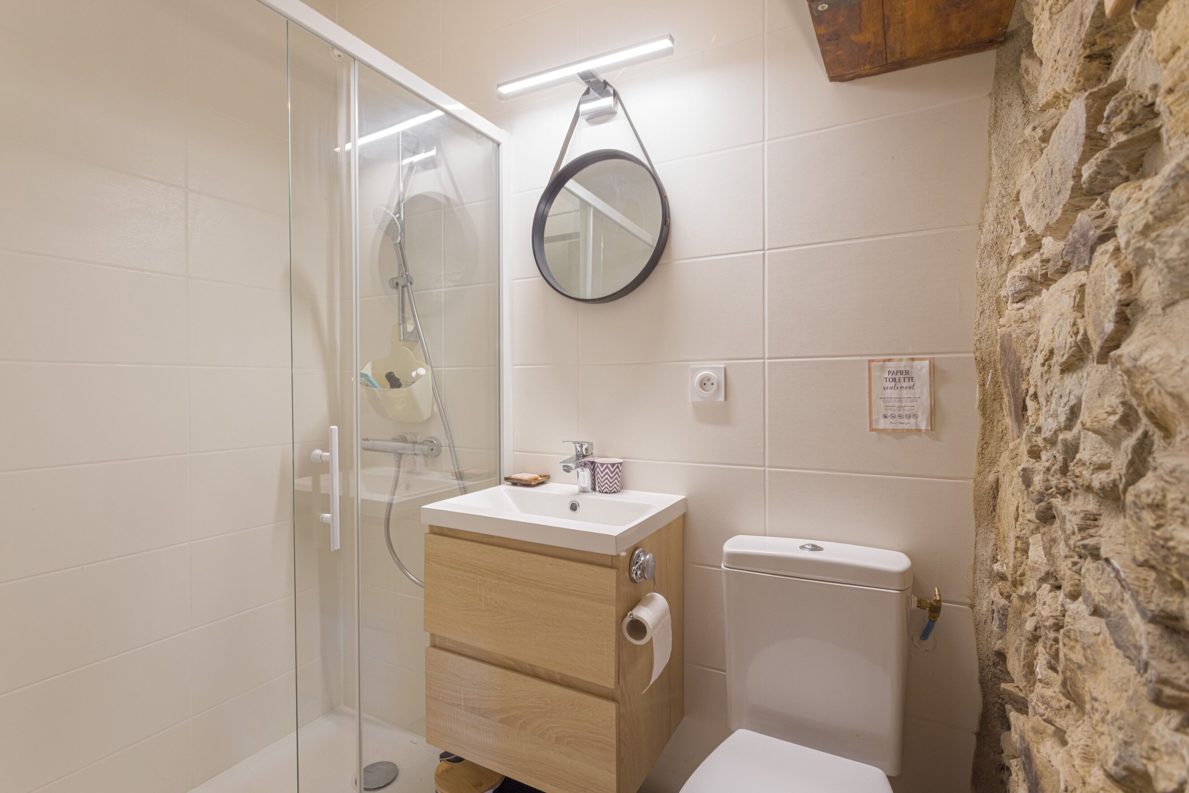 Une salle de bain avec une douche à parois de verre, un meuble-lavabo en bois avec un évier, des toilettes et un mur en pierre.