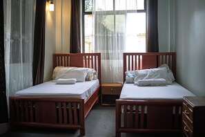 Une chambre avec deux lits en bois, chacun muni d’un oreiller et d’une couverture, une petite table de chevet en bois et une fenêtre avec des rideaux légers.