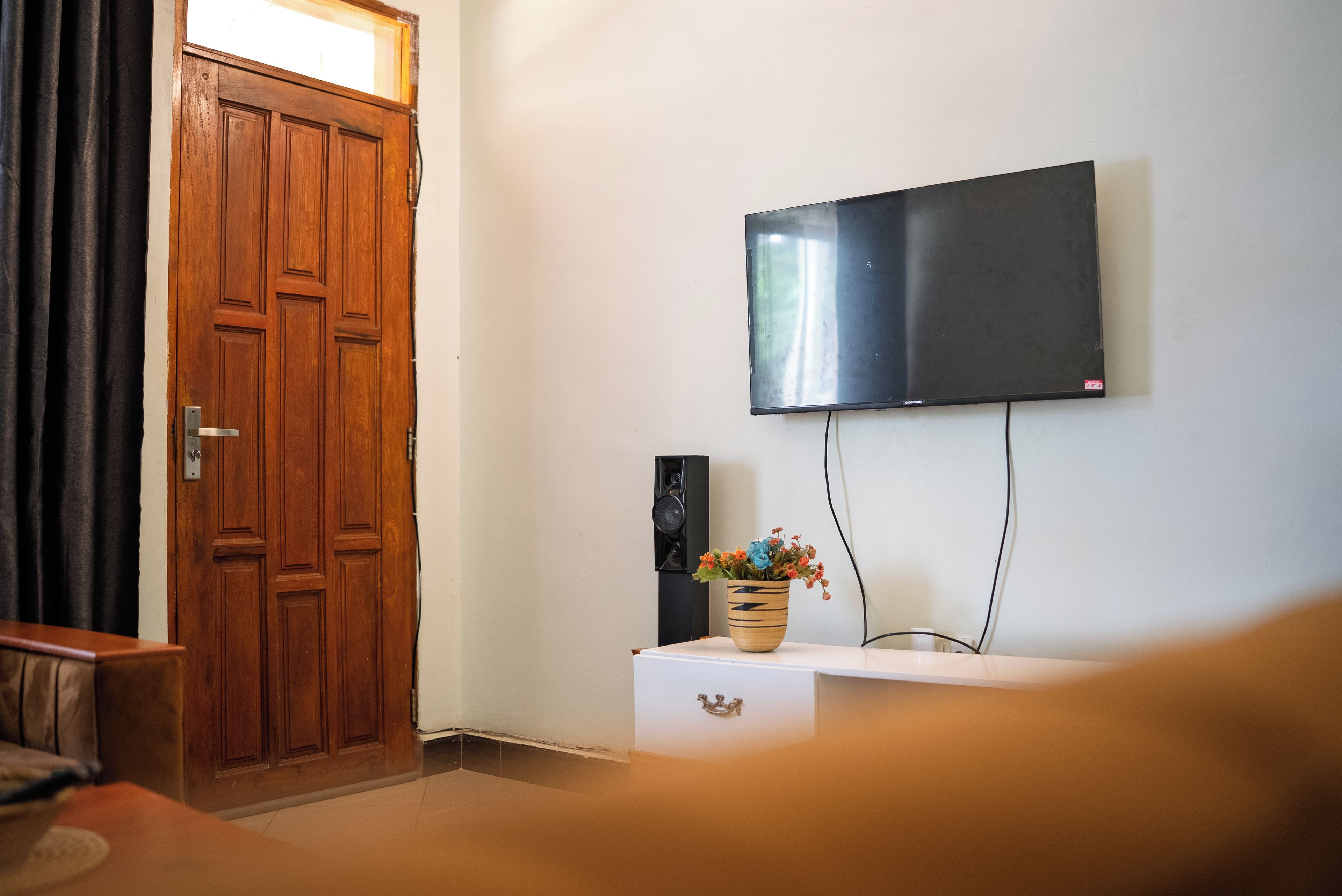 Ein Zimmer mit Holztür, einem flachbildfernseher an der Wand und einem kleinen Tisch mit Blumen in einer Vase.