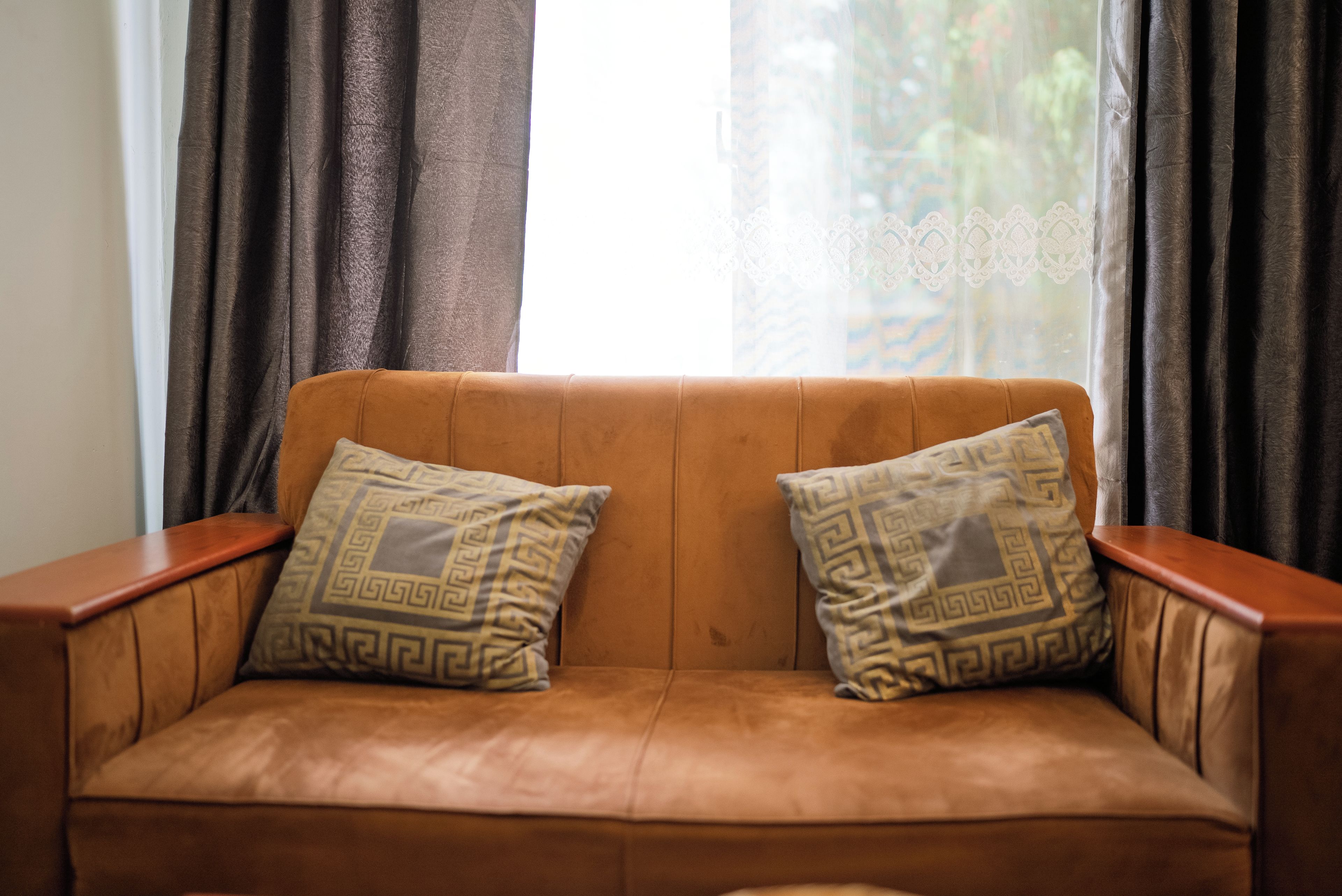 Ein braunes Leder Sofa mit zwei gemusterten Kissen, vor einem Fenster mit durchsichtigen Vorhängen platziert.