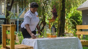 Una persona sirve comida en una mesa al aire libre con mantel blanco, rodeada de vegetación y un edificio al fondo.