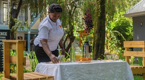 Eine Person serviert Essen an einem Tisch im Freien mit weißer Tischdecke, umgeben von viel Grün und einem Gebäude im Hintergrund.