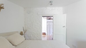 Quarto com cama, porta branca, parede de pedra e vista para outro cômodo.