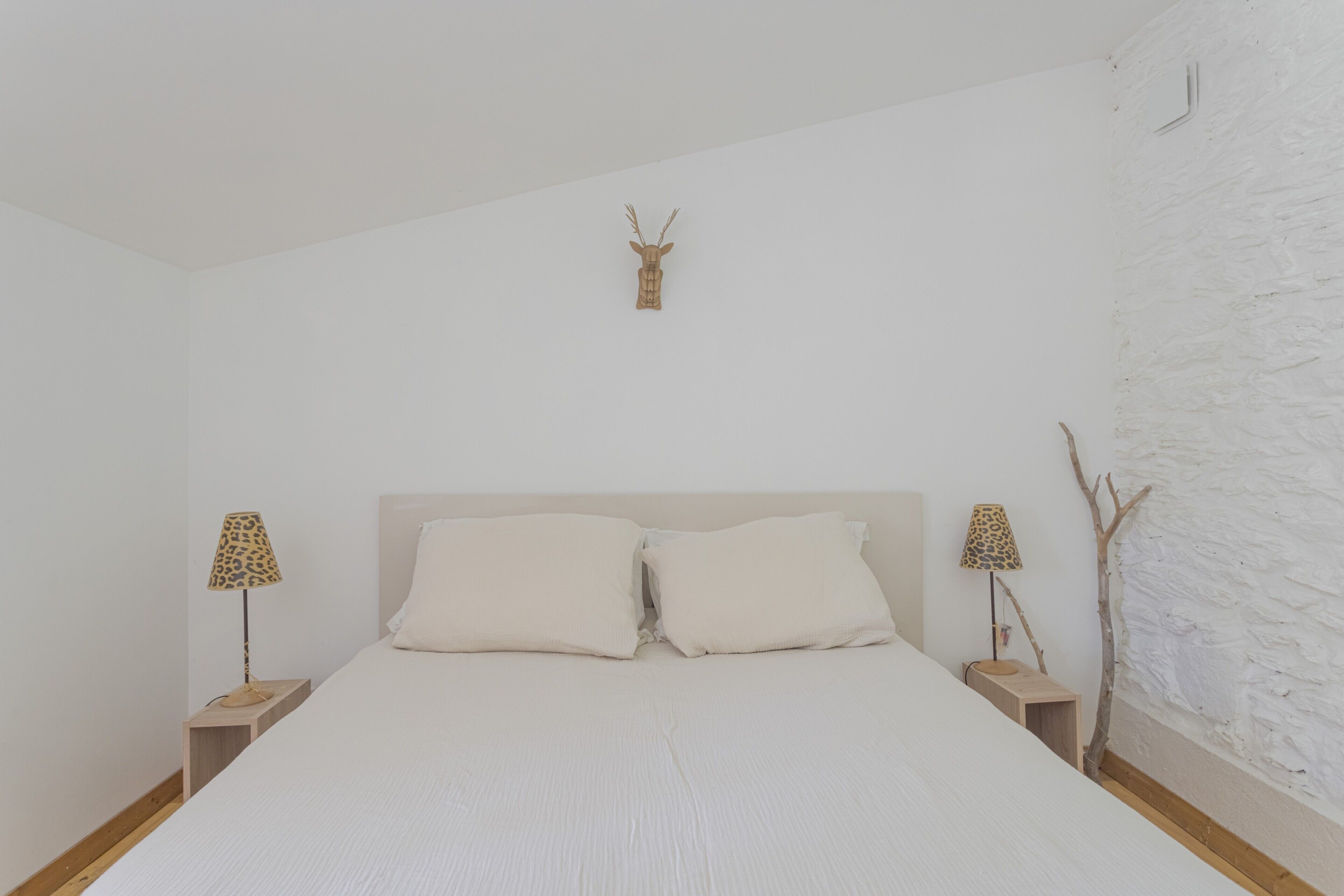 Un dormitorio con cama, dos mesitas de noche, una lámpara, una cabeza de ciervo montada en la pared y un muro con textura.