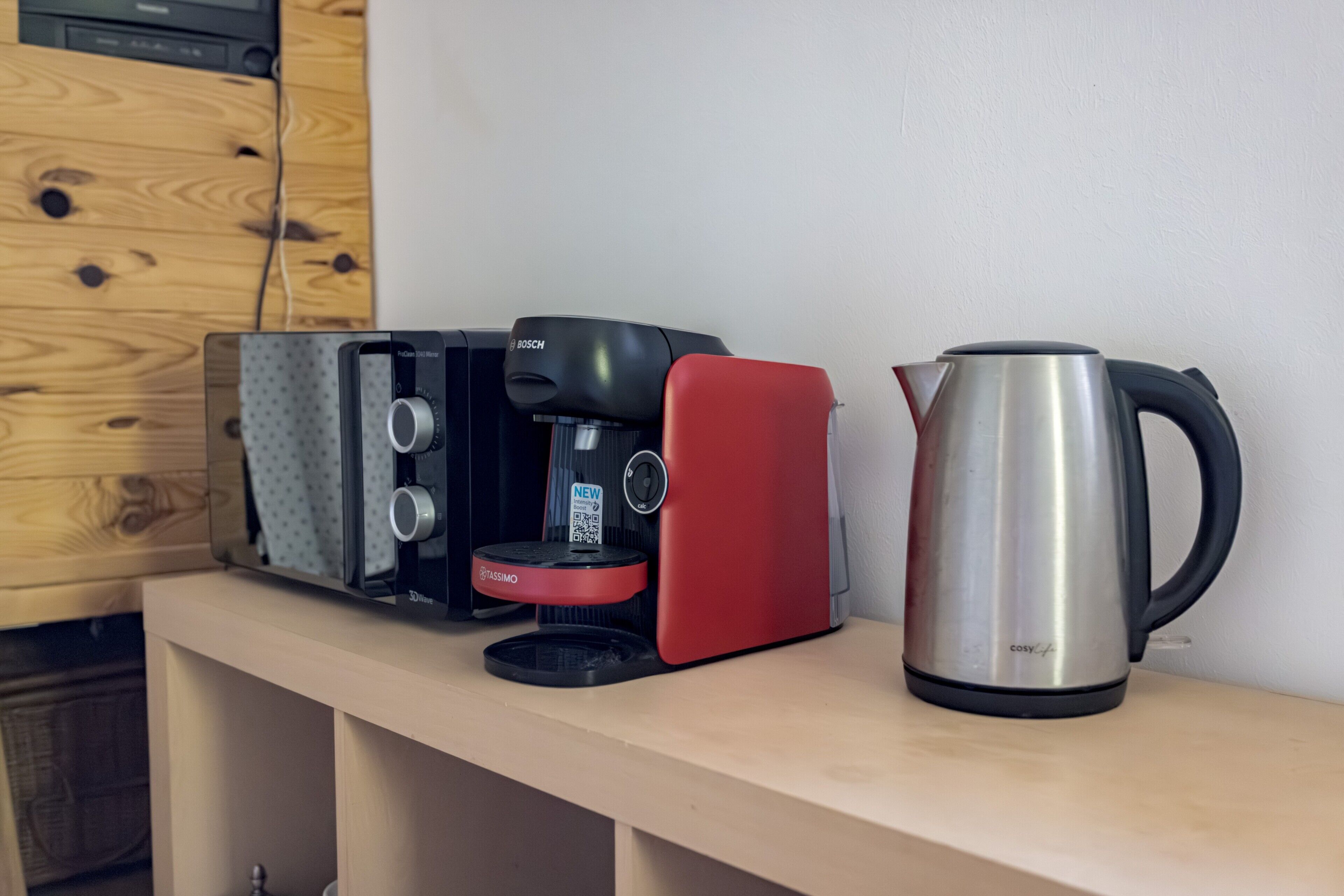 A coffee maker, a red appliance, and a stainless steel kettle on a wooden shelf.